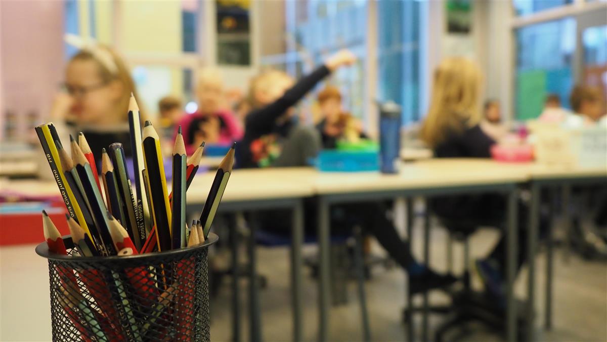 Illustrasjon fra klasserom med fokus på blyanter i forgrunnen med uklare elever bak. - Klikk for stort bilete